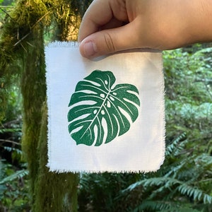 Könnte beinhalten: Ein weißes Stoffquadrat mit einem grünen Monstera-Blatt-Druck. Der Stoff hat einen ausgefransten Rand und wird von einer Hand vor einem grünen, belaubten Hintergrund gehalten.
