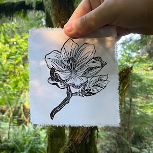 May include: A black and white print of a flower on white fabric. The fabric has a frayed edge. The print is held in front of a green forest background.