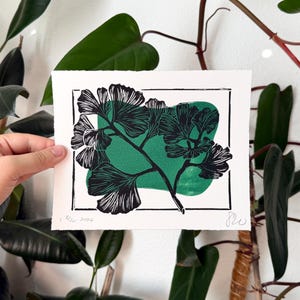 A linocut print in green and black ink on white paper of a diagonal ginkgo branch with eleven leaves extending out in three directions. there is a thin rectangle border round the branch, which the branch is obscuring in a few places. A blobby green rectangle is behind the plant inside the border. the print is held in front of green plants and a white wall.