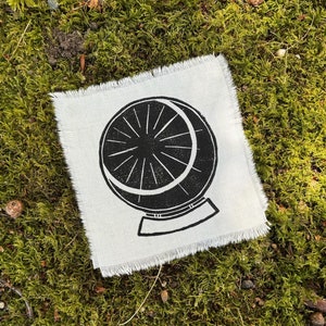 May include: A black and white printed fabric patch featuring a design of a crystal ball on a stand. The patch has a frayed edge.