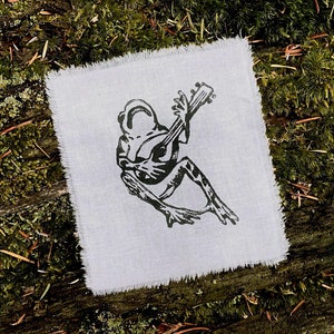 May include: A white fabric patch with a black and white illustration of a frog playing a guitar.