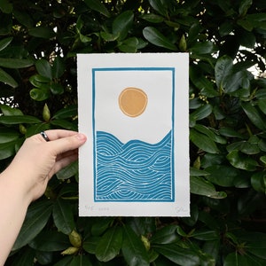medium blue and yellow linocut print showing a yellow sun on white sky over rolling blue ocean waves. the print is seven inches wide and ten inches tall and has a blue border. the waves have curved lines reminiscent of Japanese woodblock prints.