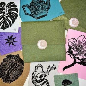 May include: A collection of handmade cards featuring various designs, including a monstera leaf, a teapot, a flower, a trilobite, a frog playing a guitar, and a small beetle. The cards are printed on different colored paper and have a rustic, handmade aesthetic.