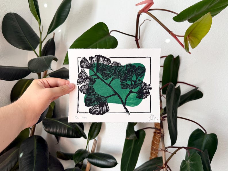A linocut print in green and black ink on white paper of a diagonal ginkgo branch with eleven leaves extending out in three directions. there is a thin rectangle border round the branch, which the branch is obscuring in a few places. A blobby green rectangle is behind the plant inside the border. the print is held in front of green plants and a white wall.