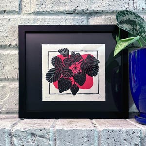 The print framed in a black frame and sitting on a white brick mantle. A green pothos plant in a dark blue pot sits next to the frame.