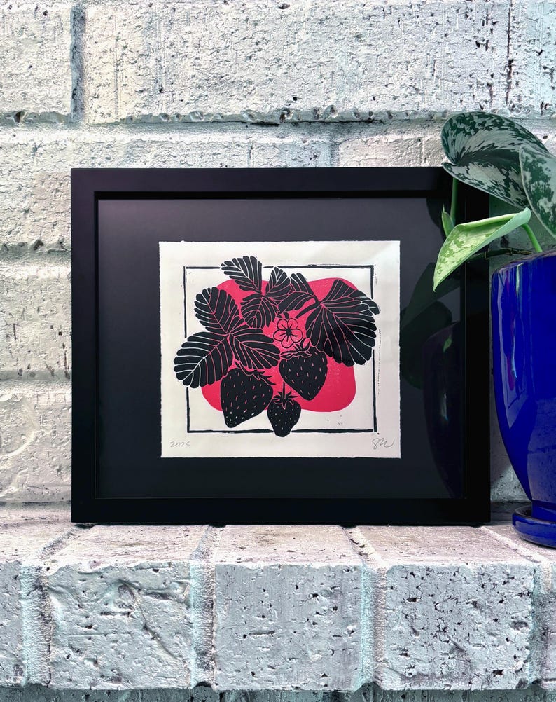 The print framed in a black frame and sitting on a white brick mantle. A green pothos plant in a dark blue pot sits next to the frame.