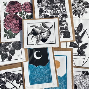 variety of shop's prints. a lemon branch, ginkgo, dahlia, dogwood, strawberries, rubber plant, a large vertical print of small houses, two prints with a sun and moon over blue ocean waves, and a rose print with green leaves and dark pink roses.