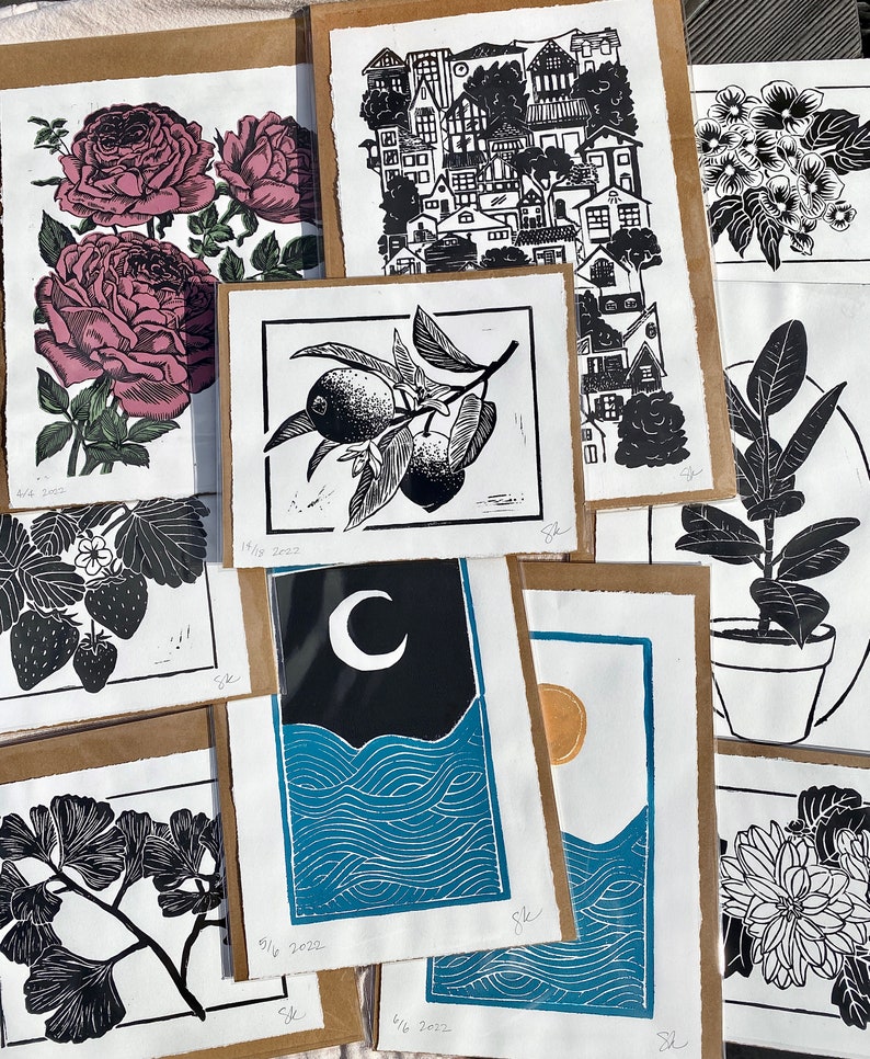 variety of shop's prints. a lemon branch, ginkgo, dahlia, dogwood, strawberries, rubber plant, a large vertical print of small houses, two prints with a sun and moon over blue ocean waves, and a rose print with green leaves and dark pink roses.
