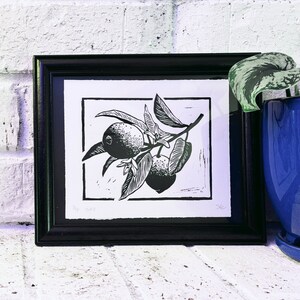 The print framed in a black frame and sitting on a white brick mantle. A green pothos plant in a dark blue pot sits next to the frame.