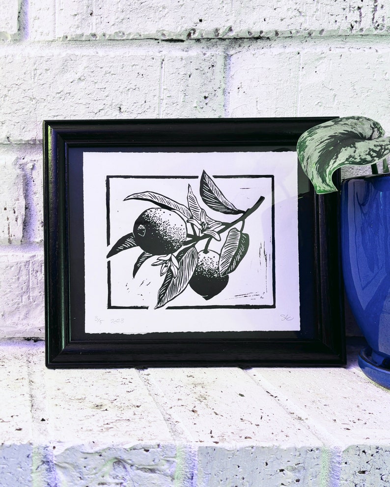 The print framed in a black frame and sitting on a white brick mantle. A green pothos plant in a dark blue pot sits next to the frame.