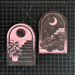 The rubber stamps used to make this print. The two blocks show the reverse images of the day and night archways. They are positioned on a cutting mat with half-inch lines going vertically and horizontally.