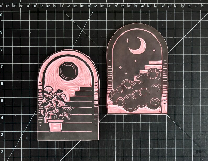 The rubber stamps used to make this print. The two blocks show the reverse images of the day and night archways. They are positioned on a cutting mat with half-inch lines going vertically and horizontally.