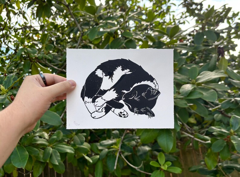 The same cat print in its entirety, viewed straight on held in front of a green rhododendron bush.