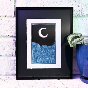 The print framed in a black frame and sitting on a white brick mantle. A green pothos plant in a dark blue pot sits next to the frame.