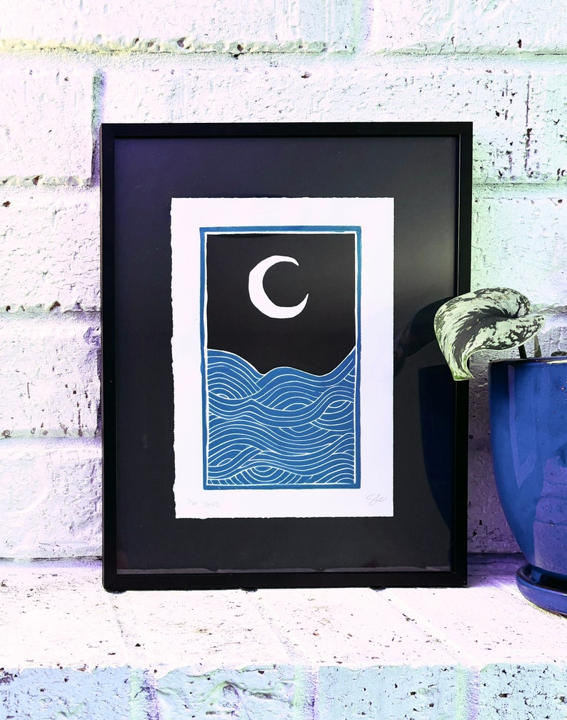 The print framed in a black frame and sitting on a white brick mantle. A green pothos plant in a dark blue pot sits next to the frame.