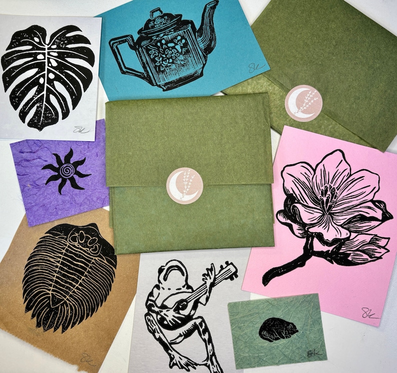 May include: A collection of handmade cards featuring various designs, including a monstera leaf, a teapot, a flower, a trilobite, a frog playing a guitar, and a small beetle. The cards are printed on different colored paper and have a rustic, handmade aesthetic.