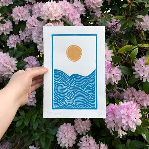 The print held in front of a blooming pink and dark green rhododendron bush