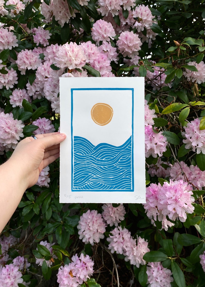 The print held in front of a blooming pink and dark green rhododendron bush