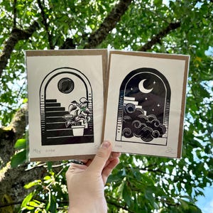 The two prints held in one hand in front of green tree branches. Each print is packaged in a plastic sleeve with a piece of cardboard behind it.