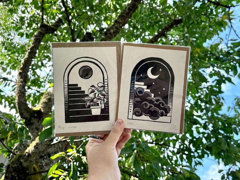 The two prints held in one hand in front of green tree branches. Each print is packaged in a plastic sleeve with a piece of cardboard behind it.