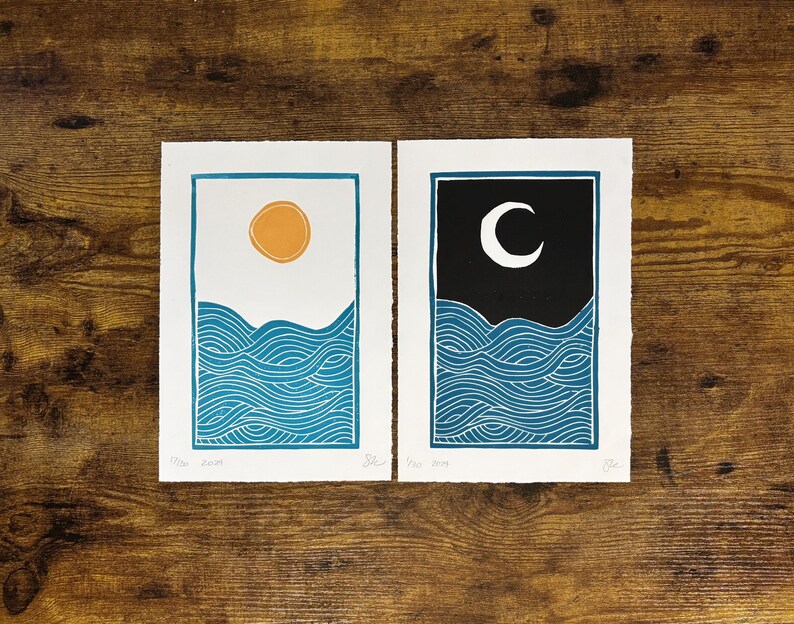 This print on a wooden desk top next to its companion print, which has the same dimensions and ocean wave design with a darker blue color. It has a black sky and white moon instead of a white sky and yellow sun.