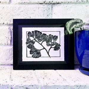 The print in a black 8 by 10 frame on a white brick mantle. A dark blue plant pot sits next to it, with a green plant leaf sticking out of the top.