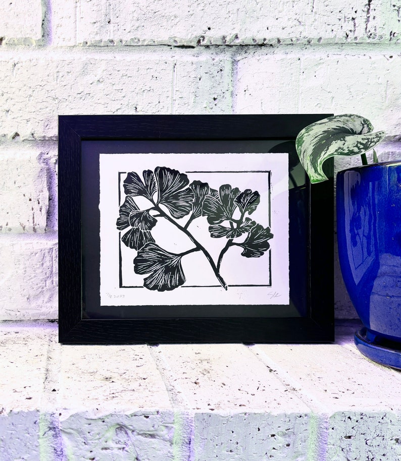 The print in a black 8 by 10 frame on a white brick mantle. A dark blue plant pot sits next to it, with a green plant leaf sticking out of the top.