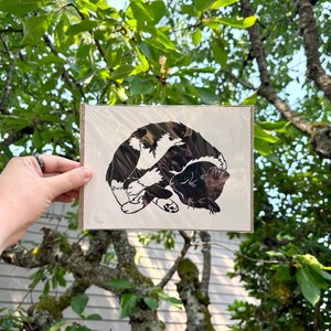 The print packaged in a clear plastic sleeve with a sheet of cardboard behind it. It is being held in front of green tree branches.