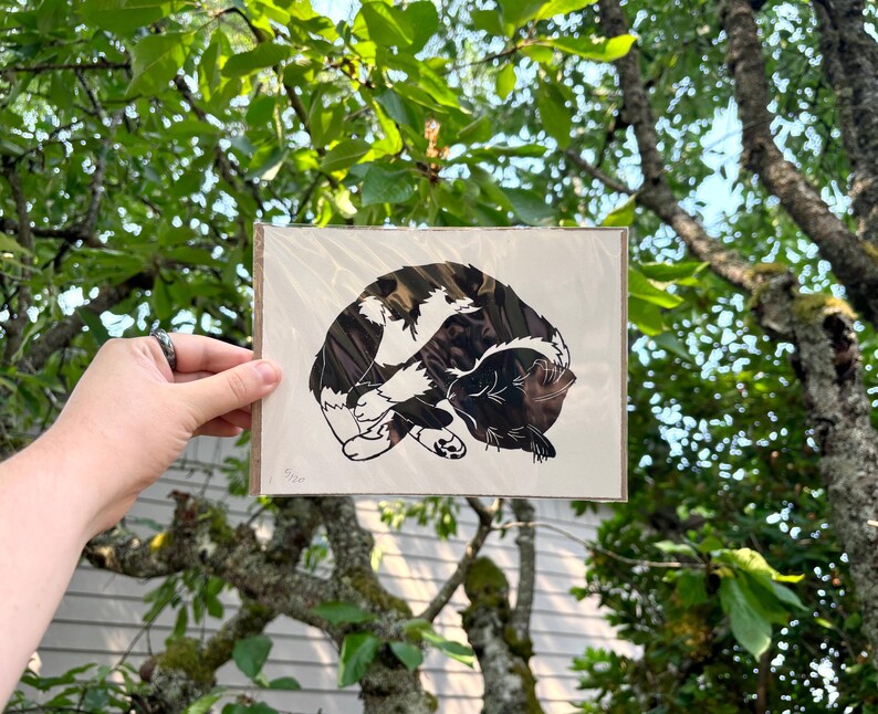 The print packaged in a clear plastic sleeve with a sheet of cardboard behind it. It is being held in front of green tree branches.