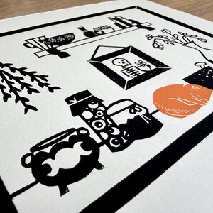 a close-up of the left side of the print. a bundle of herbs is hanging upside down on the wall next to the shelf. there are two jars next to the cat, one of which is full of eyeballs, and a small cauldron with a moon phases design on it.