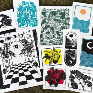 Some of the artists other prints. A pair of sun and moon over the blue ocean prints, a print with about fifty houses stacked as though on a hill, a leafy monstera, a sleeping tuxedo cat, a ginkgo branch, lemon branch, and strawberry plant. there is also a large print of a magical portal archway with an eye on it and checkerboard floor leading up to it, and two large leafy potted plants on either side.