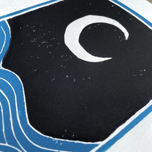 Closeup on the black sky and white moon. The crescent moon is not quite a perfect circle. The sky has speckles of white showing through, looking like stars. The closeup shows a slight rough texture of the ink on the white paper.