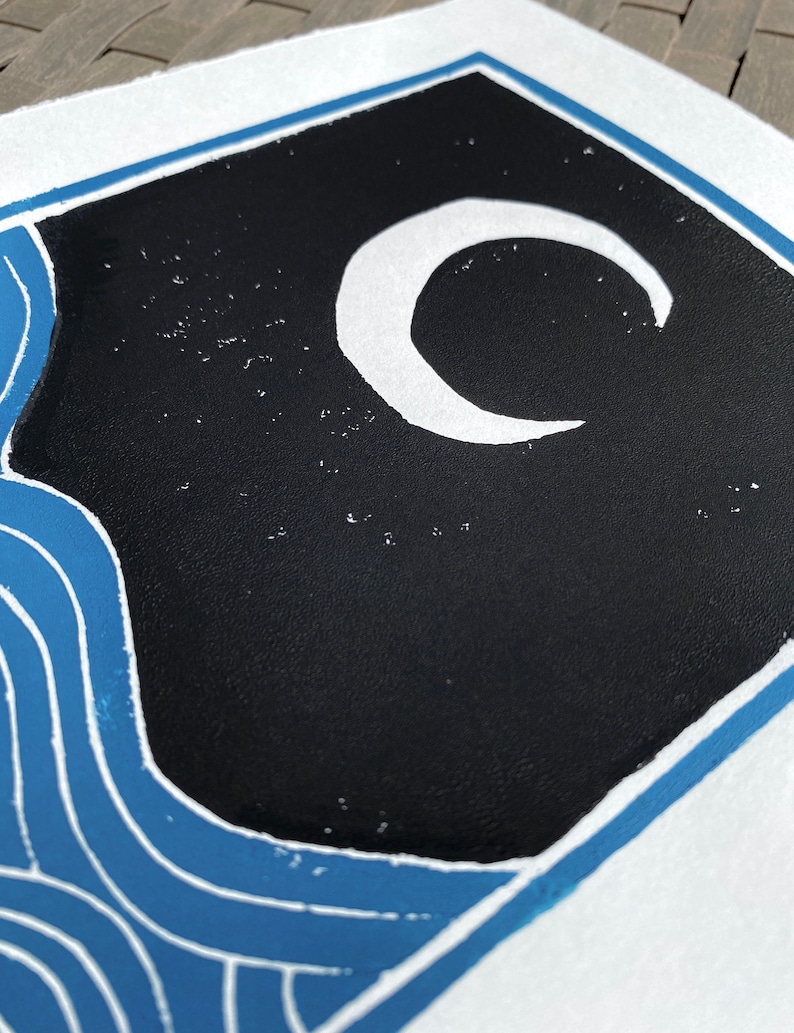 Closeup on the black sky and white moon. The crescent moon is not quite a perfect circle. The sky has speckles of white showing through, looking like stars. The closeup shows a slight rough texture of the ink on the white paper.
