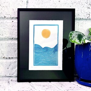 The print framed in a black frame and sitting on a white brick mantle. A green pothos plant in a dark blue pot sits next to the frame.
