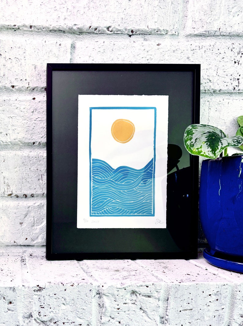The print framed in a black frame and sitting on a white brick mantle. A green pothos plant in a dark blue pot sits next to the frame.
