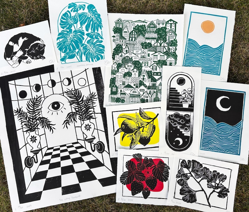 Some of the artists other prints. A pair of sun and moon over the blue ocean prints, a print with about fifty houses stacked as though on a hill, a leafy monstera, a sleeping tuxedo cat, a ginkgo branch, lemon branch, and strawberry plant. there is also a large print of a magical portal archway with an eye on it and checkerboard floor leading up to it, and two large leafy potted plants on either side.