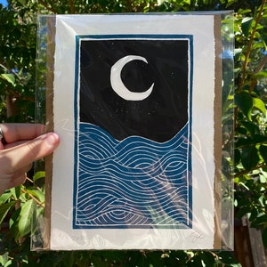 The print packaged on a piece of cardboard in a plastic sleeve for protection. The package is held in dappled shade against a green leafy background.