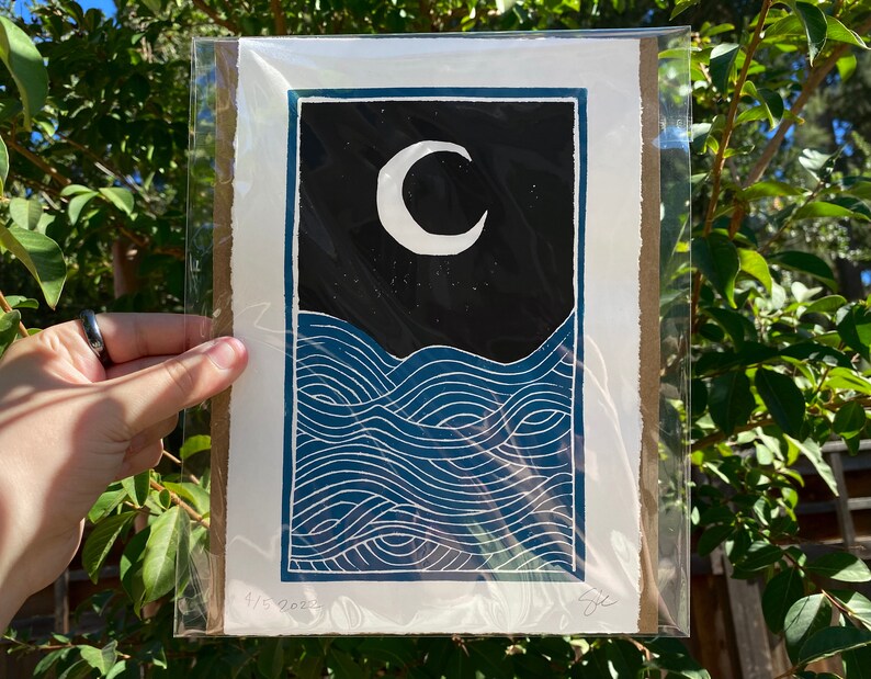 The print packaged on a piece of cardboard in a plastic sleeve for protection. The package is held in dappled shade against a green leafy background.
