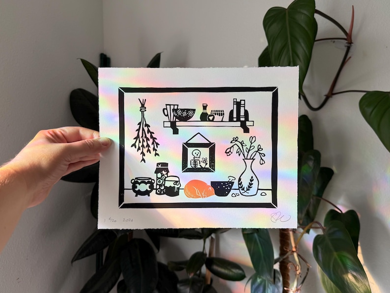 linocut print of a witch's countertop workspace. a portrait of a skeleton hangs on the wall. a jar of eyeballs and other ingredient containers line the counter, with vase of dead flowers and a sleeping orange cat in the center. on a shelf above the counter are more bottles and books. drying herbs hang down the left side of the print. theres a black border around the print edge, same as the skeleton portrait frame. Faint rainbows of light are splashed across the print from the window next to it.