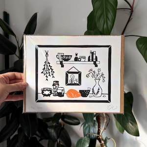 the print packaged on cardboard in a plastic sleeve. it is held in front of leafy green plants and a white wall.