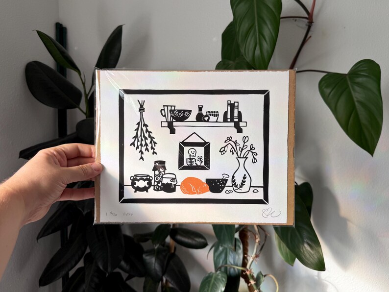the print packaged on cardboard in a plastic sleeve. it is held in front of leafy green plants and a white wall.