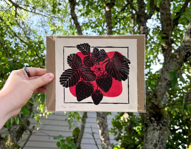 the print packaged on cardboard in a sealed plastic sleeve. the print is held in front of a lush tree