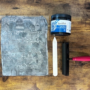 The linoleum block, ink, and rollers used to make this print. The block is a reverse image of the print. The block is glued onto a sheet of clear acrylic plastic for stability.