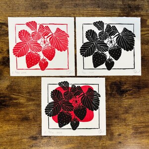 three versions of the strawberry print laying on a wooden desk. the black version is in the top right, and next to it is a red version of the same print. below them is a version of the print with a red color block blob and the black strawberries in front of it.