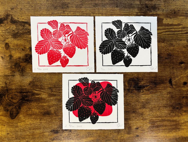 three versions of the strawberry print laying on a wooden desk. the black version is in the top right, and next to it is a red version of the same print. below them is a version of the print with a red color block blob and the black strawberries in front of it.