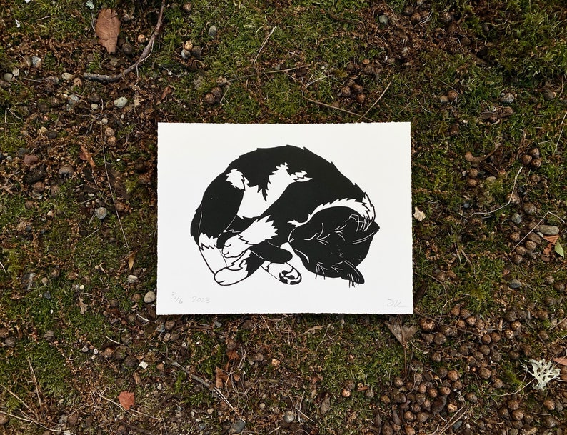 A horizontal black and white linocut print of a tuxedo cat sleeping. The cat has a black head and back with white belly and paws. It is laying on its left side with its four paws crossed over each other in front of its belly.