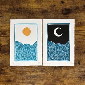 This print on a wooden desk top next to its companion print, which has the same dimensions and ocean wave design with a darker blue color. It has a black sky and white moon instead of a white sky and yellow sun.