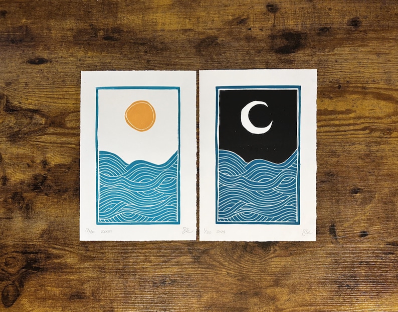 This print on a wooden desk top next to its companion print, which has the same dimensions and ocean wave design with a darker blue color. It has a black sky and white moon instead of a white sky and yellow sun.