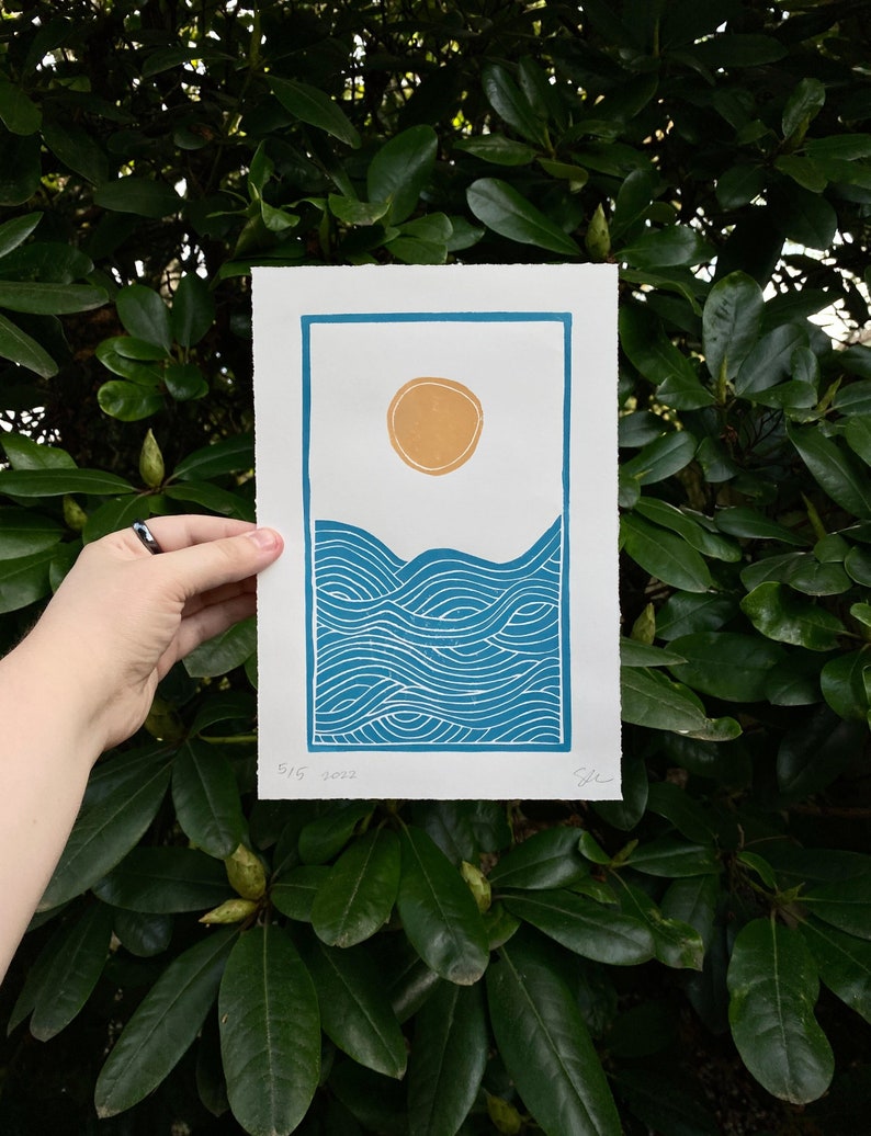 medium blue and yellow linocut print showing a yellow sun on white sky over rolling blue ocean waves. the print is seven inches wide and ten inches tall and has a blue border. the waves have curved lines reminiscent of Japanese woodblock prints.