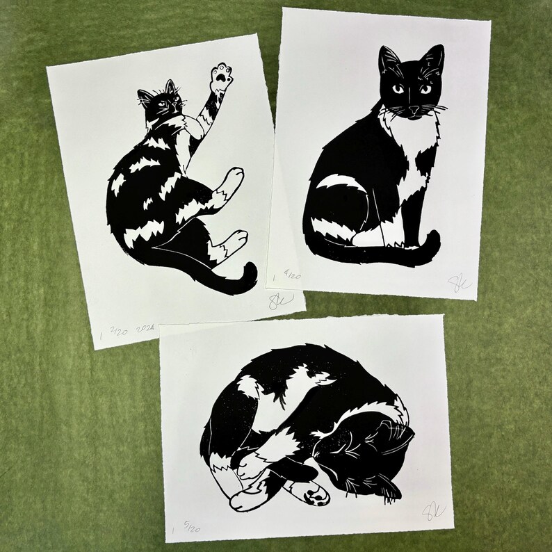 The print with two others of similar tuxedo cats. In addition to the sleeping cat, one cat is playing with its left front paw stretched upward, and the third is sitting looking at the viewer with its tail curled over its paws.